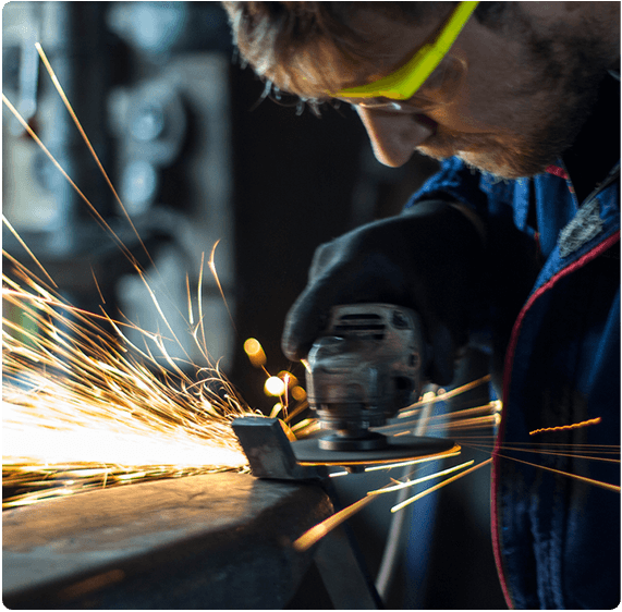A person wearing safety glasses uses a grinder on a metal piece, causing bright sparks to fly. Focused on their work, they wear dark gloves and protective clothing. The background is blurred, emphasizing the intense activity at Jumalu Fencings manufacturing site.
