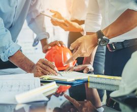A group collaborates at a table with blueprints and documents, pointing and writing. An orange construction helmet and white hard hat lie beside sketches, suggesting a manufacturers meeting on security fencing or architectural planning.