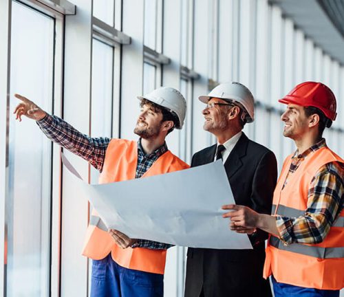 Three men in hard hats and orange safety vests stand in a modern building, looking out the window. One points while another holds a blueprint, likely discussing construction or architectural plans for the Clearview project.
