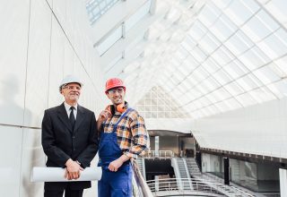 Two construction professionals stand inside a modern building with large glass panels. One, wearing a suit and gray hard hat, holds blueprints while discussing plans for a clear view fence. The other, in overalls and a red hard hat, smiles in the bright, spacious atrium.