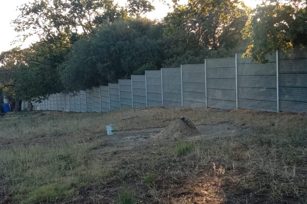 A long, gray ClamberPrufe Clear View Fencing panel runs parallel to a patch of dry, grassy ground with a green bucket and a shovel resting on a dirt mound. Dense trees and shrubs grow behind the fence.