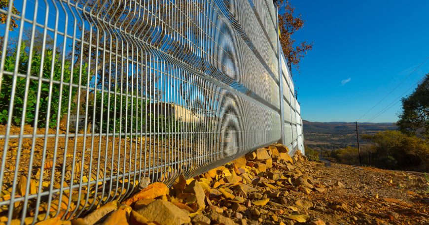 Close-up of a Jumalu fencing design with yellow and brown leaves scattered on the ground. Trees stand silhouetted against a clear blue sky, while the hilly landscape stretches out in the distance, offering a serene clearview.