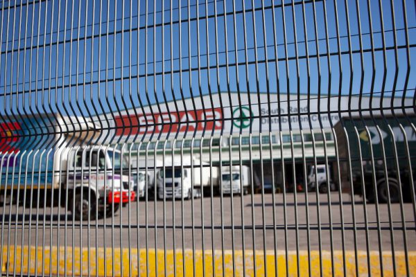 Peering through a wire fence into the logistics facility, several trucks stand idle before the warehouse, adorned with manufacturer logos. The sky is a clearview blue, with the fence casting wavy distortions over this vibrant scene.