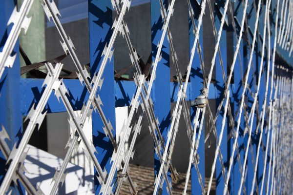 Close-up of a blue metal security fence with an interlocking razor wire pattern. The sharp, silver blades crisscross against a blurred background of buildings and sky. Bright sunlight casts shadows, highlighting the Jumalu fencing’s structure.