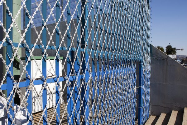 The blue and white chain-link fence, a product of a top security fencing manufacturer, features barbed wire atop at an industrial site. Sunlight casts shadows on the concrete below, while trees and a distant horizon enhance the backdrop.