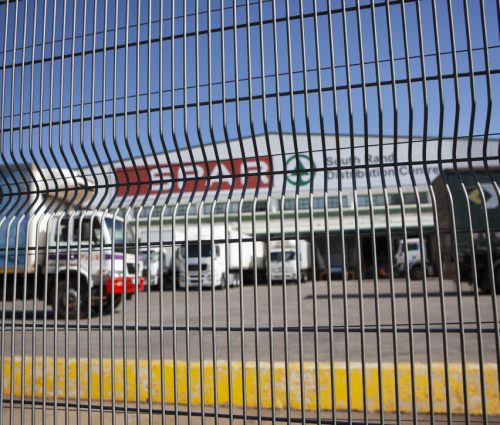 Behind the tall jumalu fencing with its distinctive wavy pattern, a commercial loading area comes into view. Trucks and vans are subtly distorted by the design, while a building stands in the background under a bright blue sky.