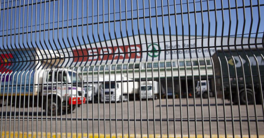Behind the tall jumalu fencing with its distinctive wavy pattern, a commercial loading area comes into view. Trucks and vans are subtly distorted by the design, while a building stands in the background under a bright blue sky.