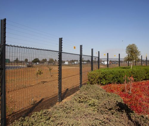 A clear view fence stretches alongside a road with a barren field on one side and a landscaped area featuring green and red foliage offering a clear view on the other. Trees and vehicles can be seen in the background under the pristine blue sky.