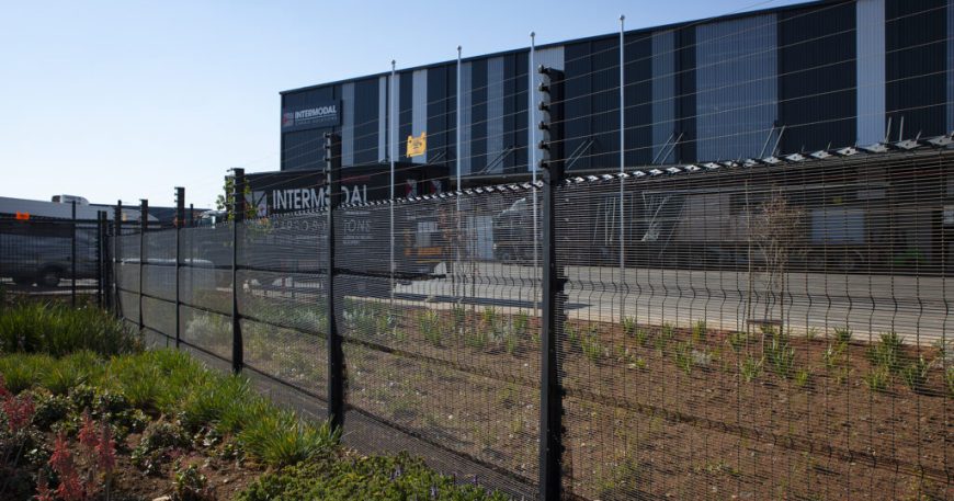 A logistics or warehouse facility with a clearview black fence in front, set against a clear blue sky. The building boasts a modern design and features a sign that reads INTERMODAL, while lush vegetation flourishes in the foreground.