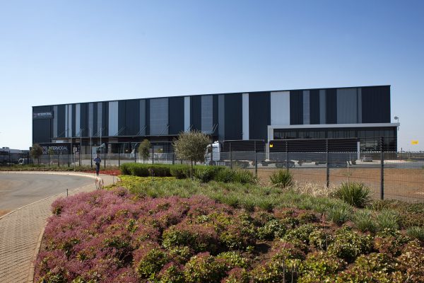 A large, modern industrial building with dark vertical panels stands under a clear blue sky. In the foreground, pink flowers and green shrubs flourish by a paved road. A clear view fence surrounds the facility, offering an unobstructed glimpse of this manufacturing marvel.