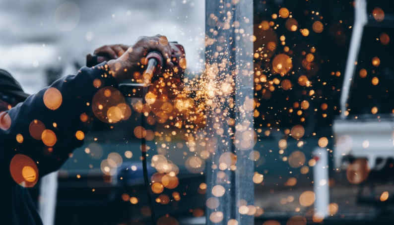 A person uses a power tool to grind a metal surface, creating a shower of bright orange and yellow sparks. The blurred background accentuates the dynamic motion and intensity of the work, offering a clear view of craftsmanship akin to the precision in crafting Jumalu fencing.