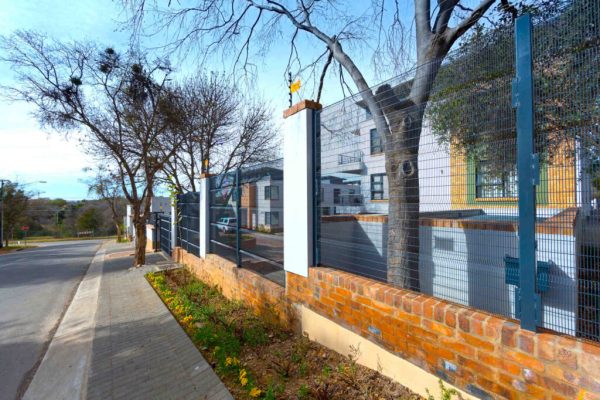 Street view of a residential area with a brick and metal fence bordering a modern house. Leafless trees line the sidewalk next to the paved road under a clear blue sky, offering a clear view. A yellow sign is mounted on the fence.
